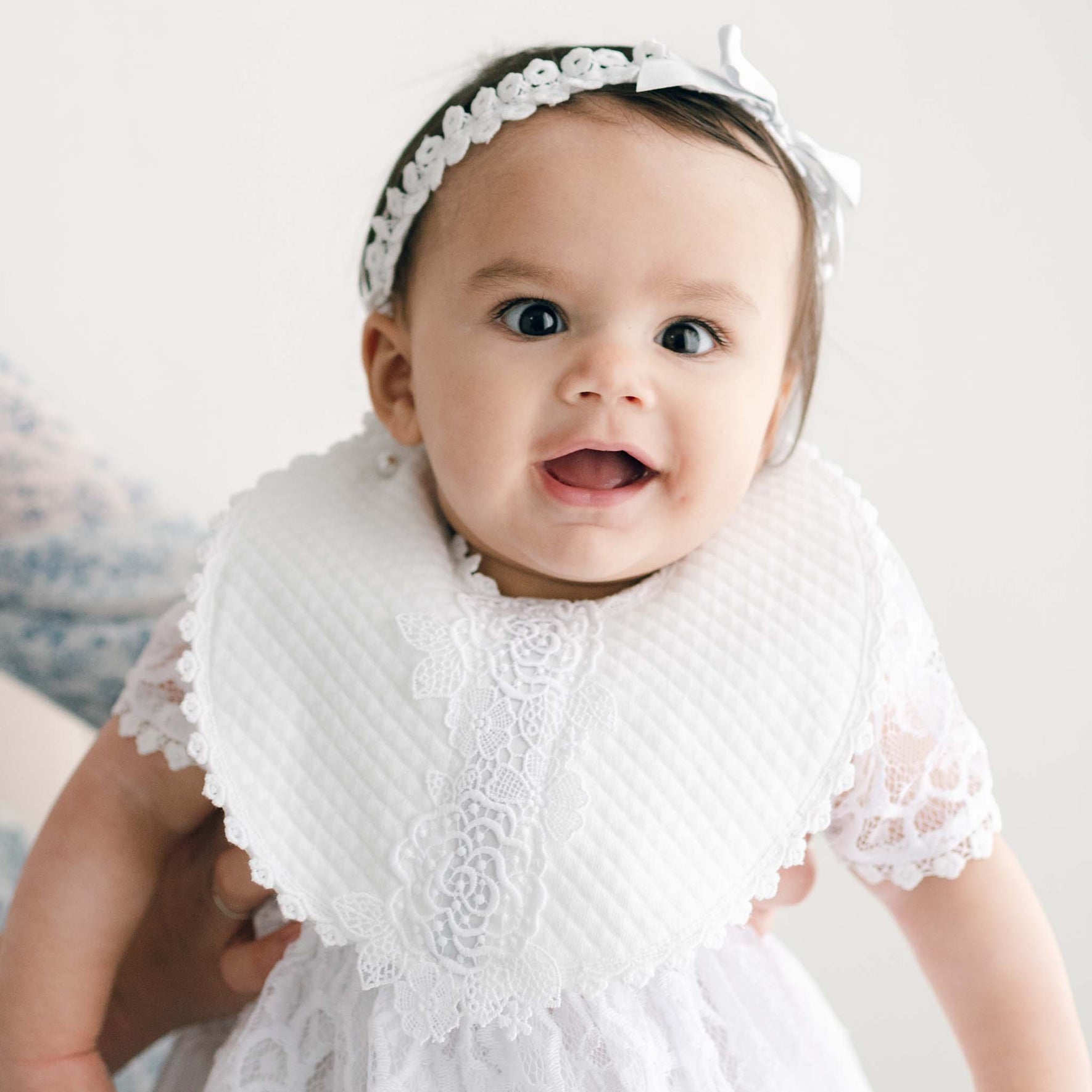 The baby is wearing a white floral lace dress and headband with a bow, smiling and looking forward. Being held by someone partially visible, the light, plain background enhances the charm of her baptism outfit featuring the Olivia Bib.
