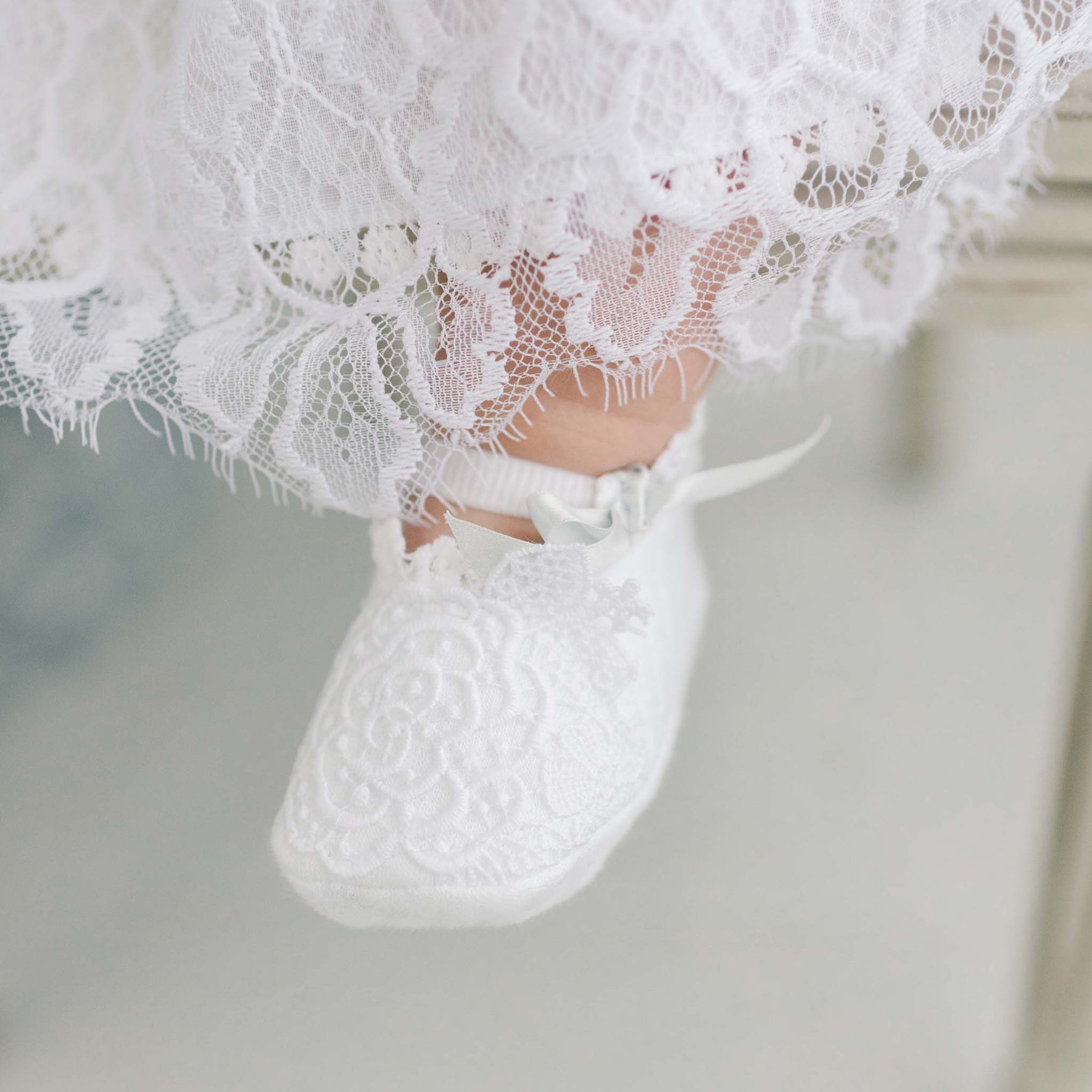 A close-up of an infant's foot wearing the Olivia Christening Booties, delicately crafted with soft white lace and enhanced by a charming floral lace applique and small pale blue silk ribbons. The hem of the baby's dress, made from matching lace fabric, is partially visible above the bootie.