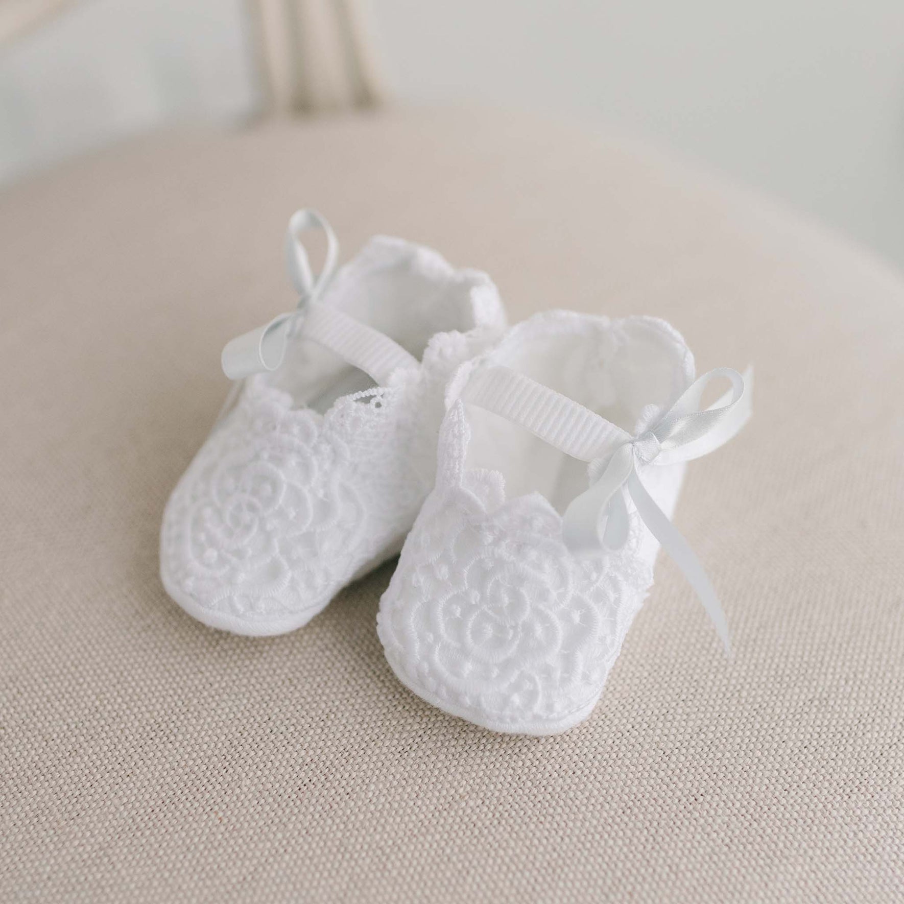 The Olivia Christening Booties, featuring intricate floral lace details and soft fabric soles, are adorned with pale blue silk ribbons and placed on a light-colored fabric surface.
