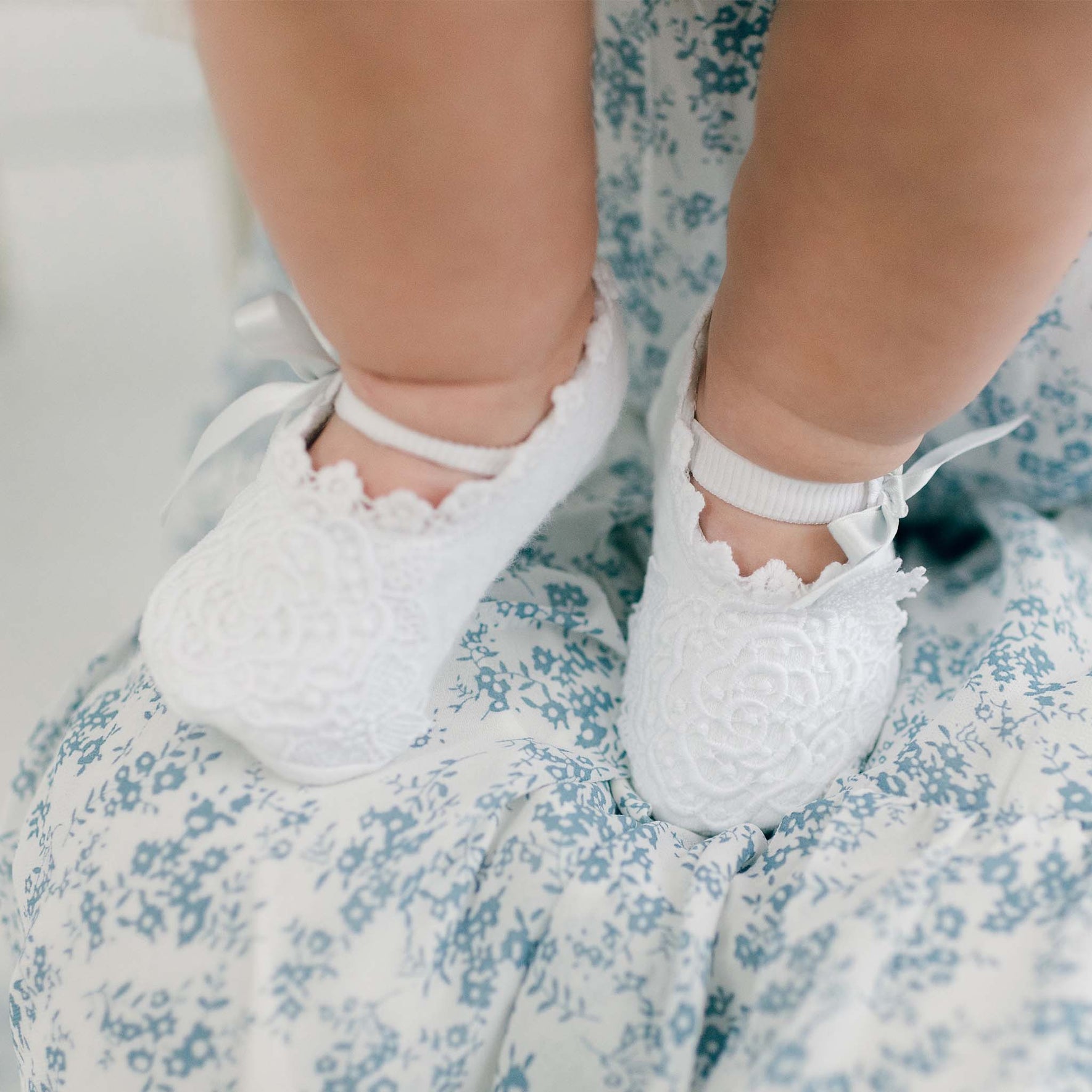 A close-up of a baby's legs and feet, adorned with the Olivia Christening Booties featuring floral lace applique. The baby is dressed in a charming floral patterned outfit in shades of blue and white, accented by delicate pale blue silk ribbons that complement the intricate details of the booties.