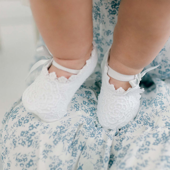 A close-up of a baby's legs and feet, adorned with the Olivia Christening Booties featuring floral lace applique. The baby is dressed in a charming floral patterned outfit in shades of blue and white, accented by delicate pale blue silk ribbons that complement the intricate details of the booties.