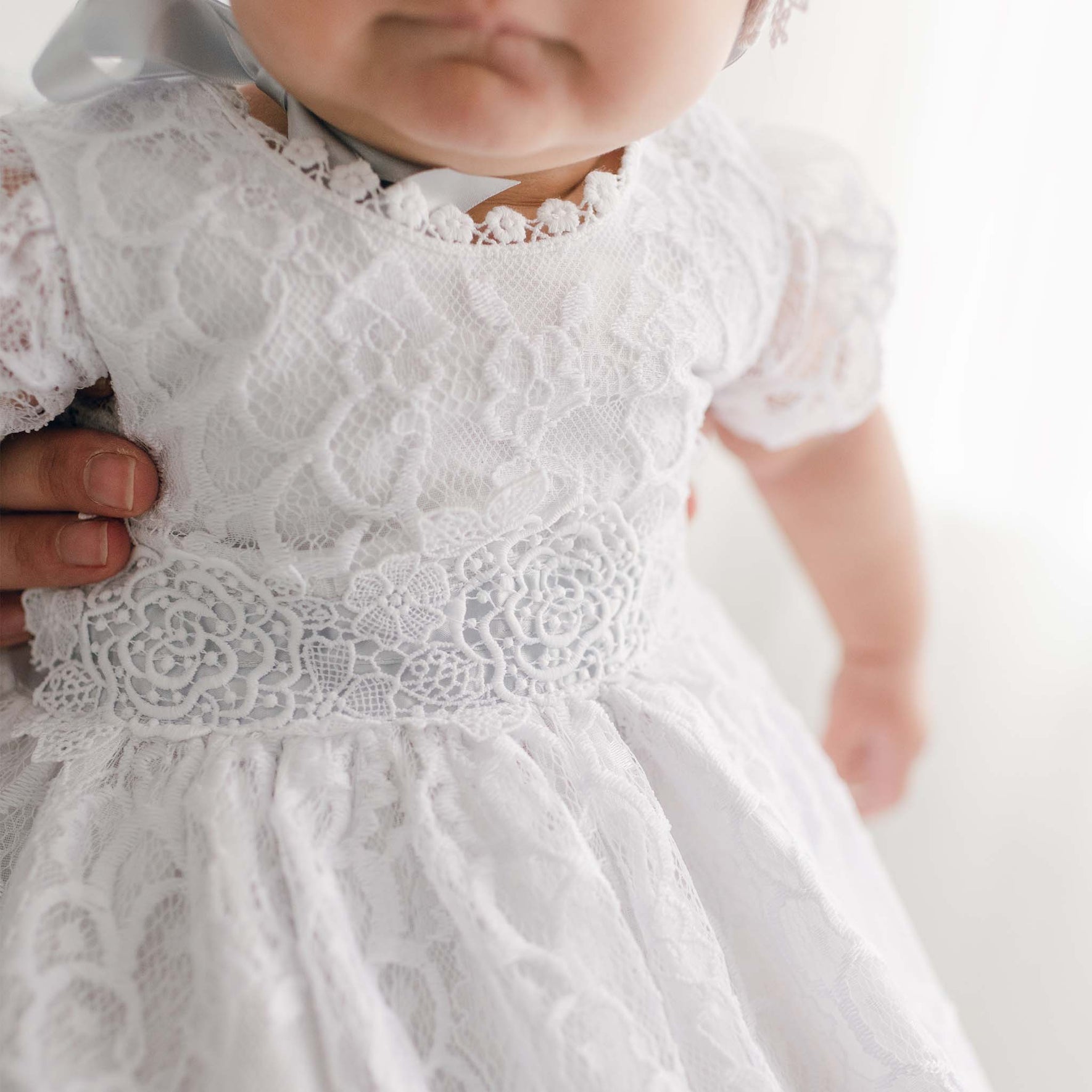 A close-up of a baby in the floral lace Olivia Dress, featuring a ribbon elegantly tied at the back. An adult's hand gently supports the child against a softly blurred background. This exquisite piece is handmade in the USA.