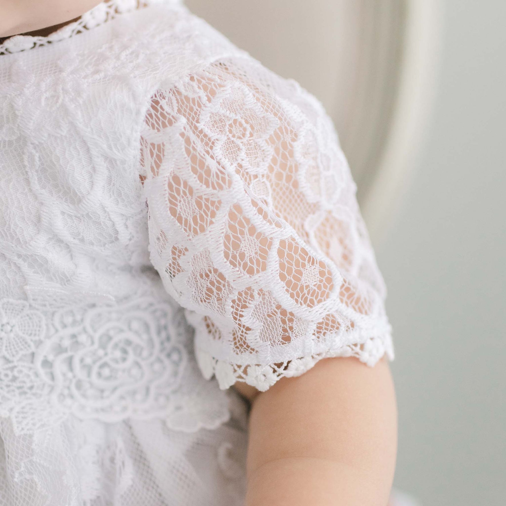 Close-up of a baby in an Olivia Dress, handmade in the USA. The emphasis is on the detailed floral lace pattern on the sleeve and bodice, elegantly highlighting part of the baby's arm and shoulder against a gentle background.