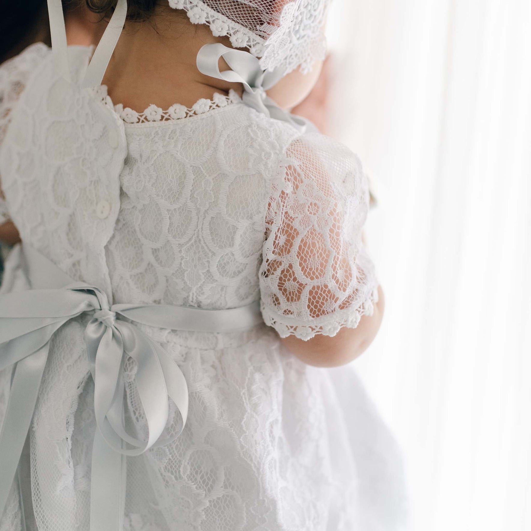 A child stands near a sheer curtain, adorned in the exquisite Olivia Dress, a handmade creation from the USA featuring intricate floral lace patterns and a bow on the back. The delicate charm is enhanced by short sleeves and a matching lace bonnet as the baby faces away.