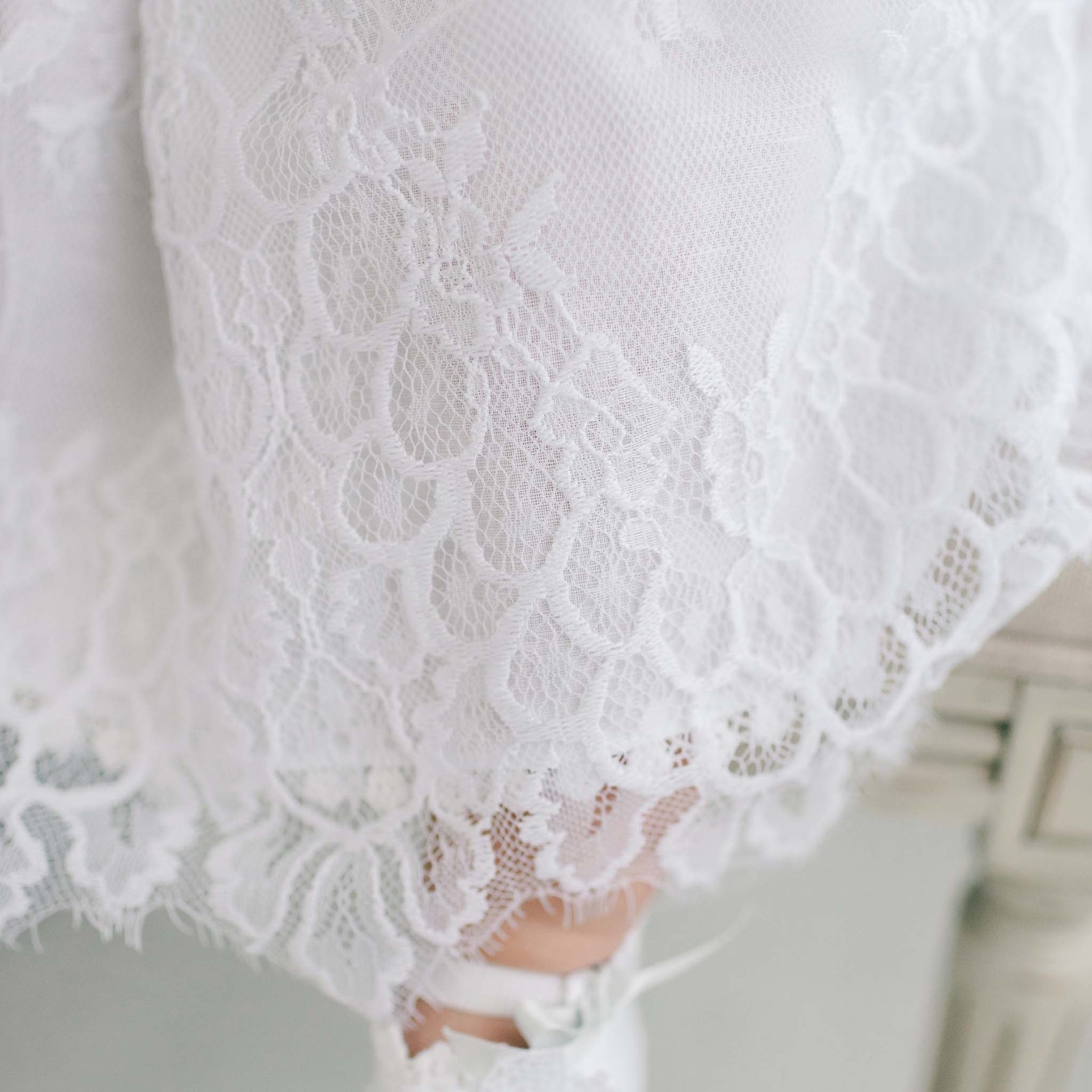 A close-up of the white floral lace fabric from the Olivia Dress, gracefully draped over a surface. The exquisite handmade details, crafted in the USA, stand out beautifully, with a satin ribbon gently emerging from the bottom right corner.