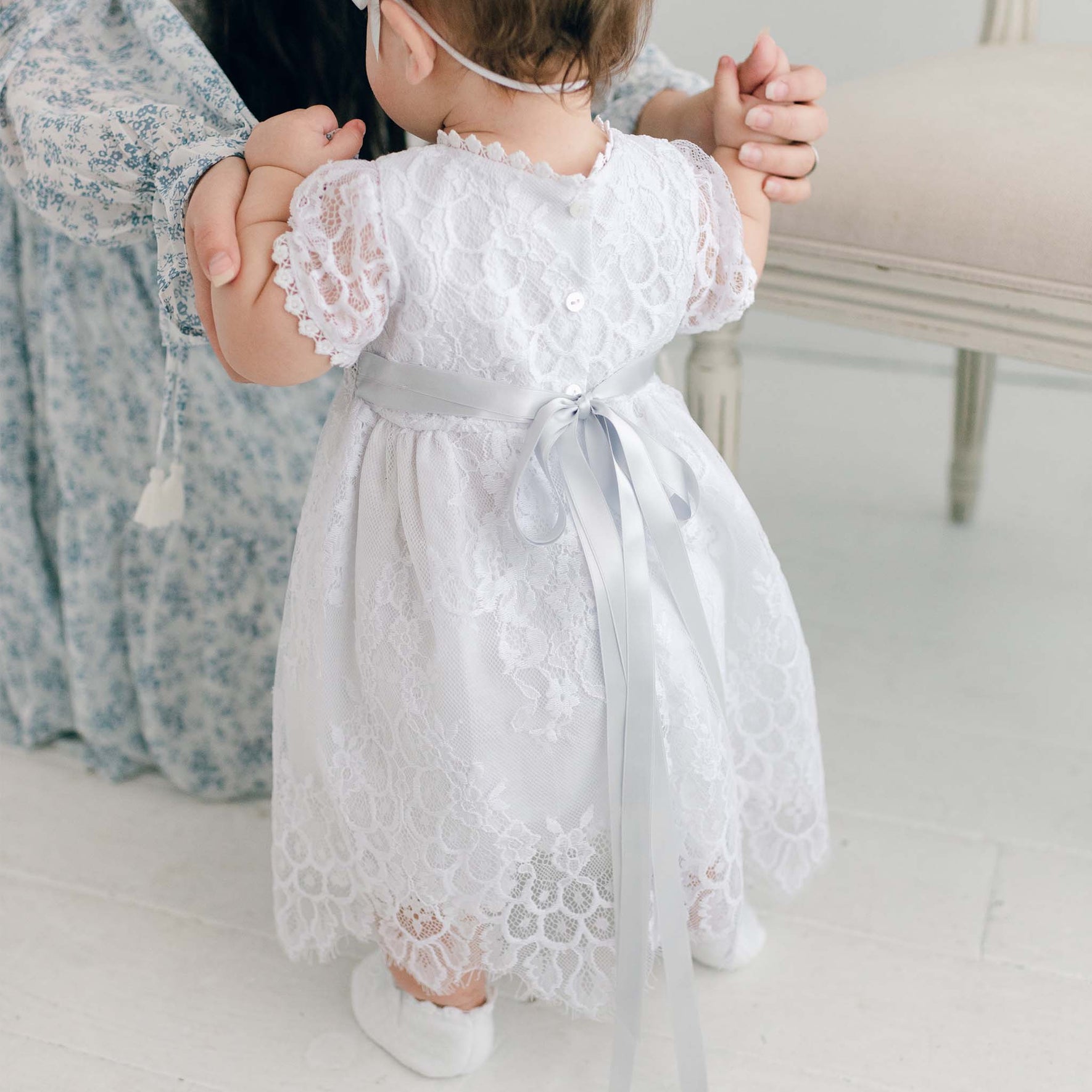 A baby wearing the Olivia Dress, crafted from floral lace and accented with a satin ribbon, stands on a white floor while holding hands with an adult dressed in a floral outfit who is kneeling. In the background, a handcrafted chair made in the USA adds an elegant touch.