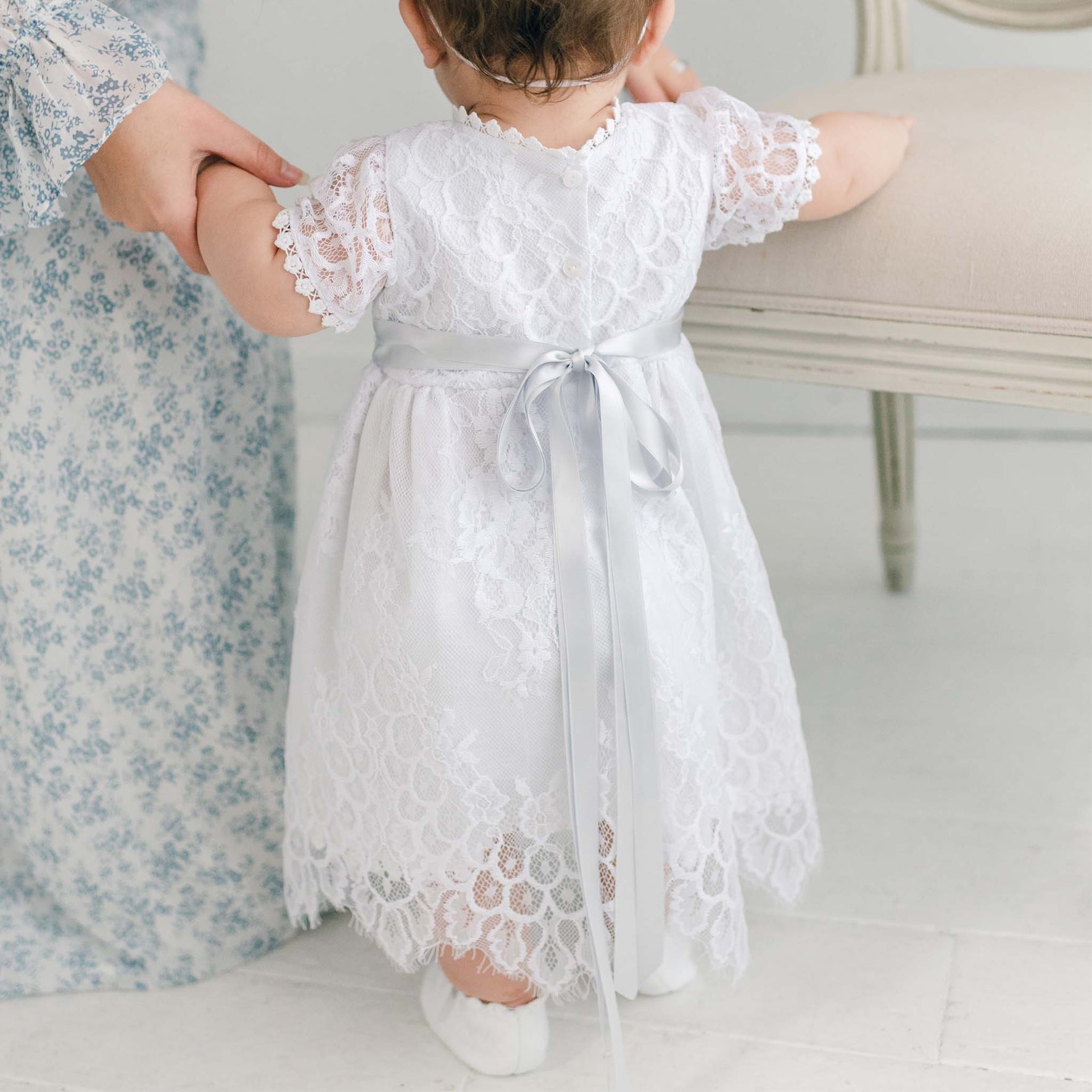 A baby stands indoors on a light-toned floor wearing a white Olivia Dress, accented with a silver ribbon, while holding an adult's hand. The adult is dressed in a floral-patterned outfit that beautifully complements the delicate design of the handmade in USA dress.