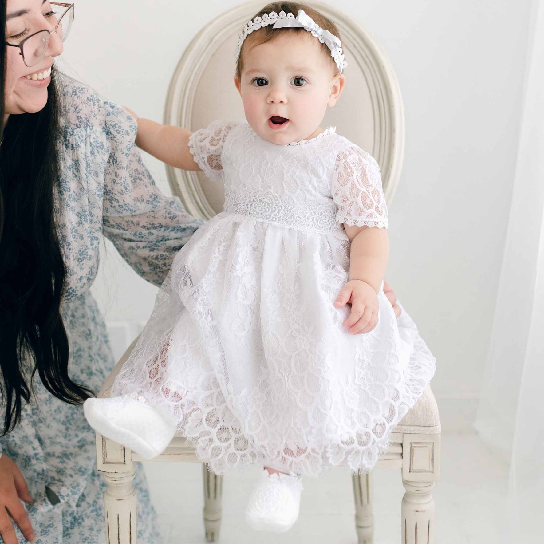 A baby wearing the Olivia Dress and a matching headband sits on a chair. The baby has white booties and gazes forward with an open mouth expression. Beside the chair, a woman with long dark hair, glasses, and a floral dress kneels down smiling.