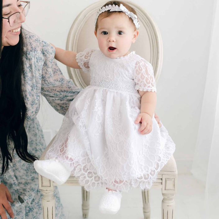 A baby wearing the Olivia Dress and a matching headband sits on a chair. The baby has white booties and gazes forward with an open mouth expression. Beside the chair, a woman with long dark hair, glasses, and a floral dress kneels down smiling.