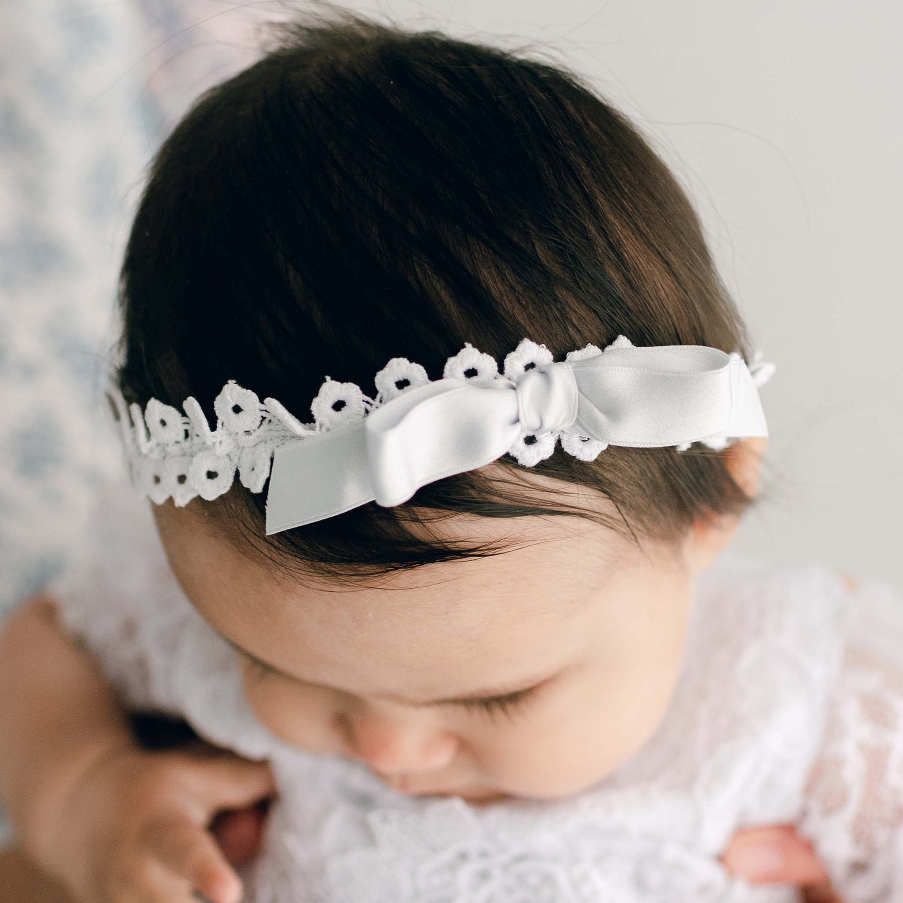 A close-up of a baby wearing the Olivia Headband, a white floral lace accessory adorned with a silk ribbon bow. Dressed in a delicate white lace outfit, the baby gazes downward.