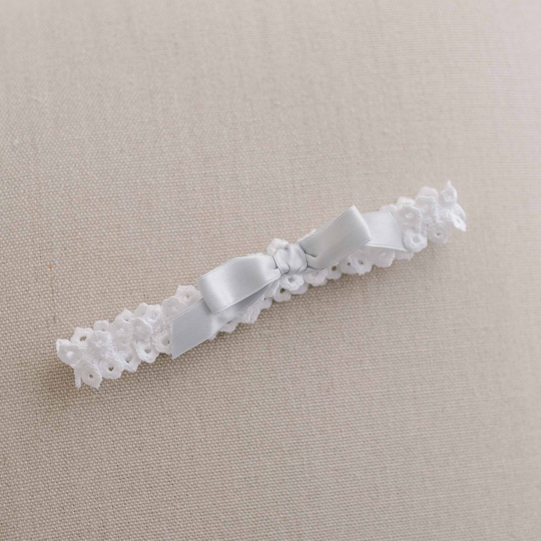 An elegant white cotton lace Olivia Headband featuring small floral patterns and a light gray silk ribbon bow is displayed on a beige fabric background.