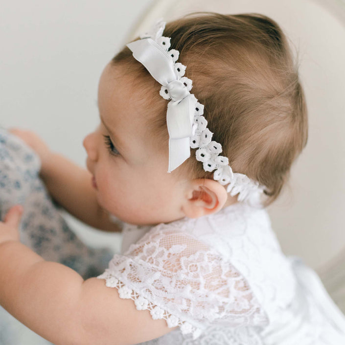 A baby is adorned in a white cotton lace dress and the Olivia Headband, featuring a delicate silk ribbon bow. Gazing softly to the side, the baby is seated in a light-colored setting, radiating innocence and charm.