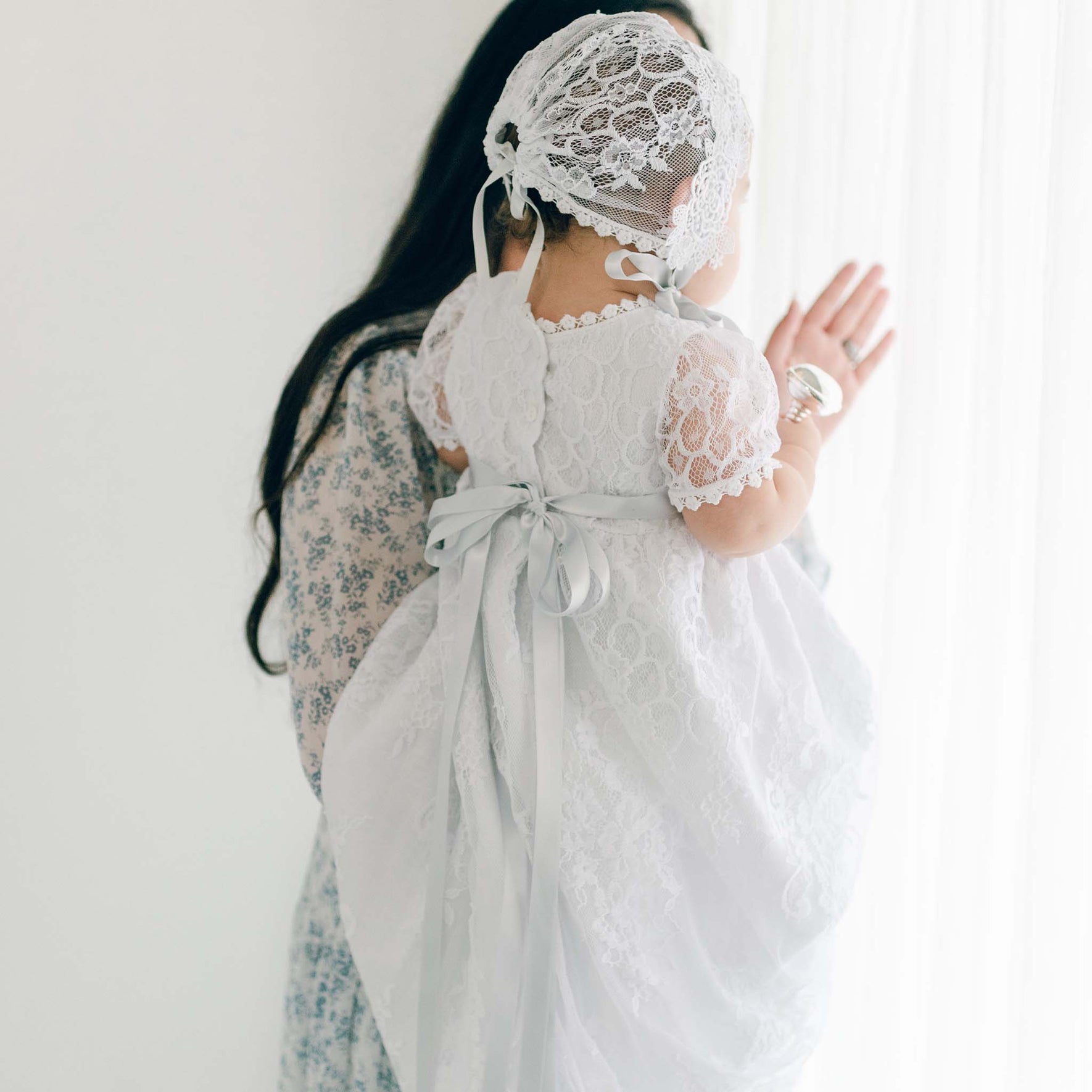A woman holds a baby dressed in a pristine white lace Christening Gown, complemented by the delicate Olivia Lace Christening Bonnet. The woman, elegantly attired in a floral-patterned dress, stands beside a window adorned with sheer curtains that cast gentle light into the room. With the baby's back turned to the camera, the intricate details of the Olivia Lace Bonnet are on full display.