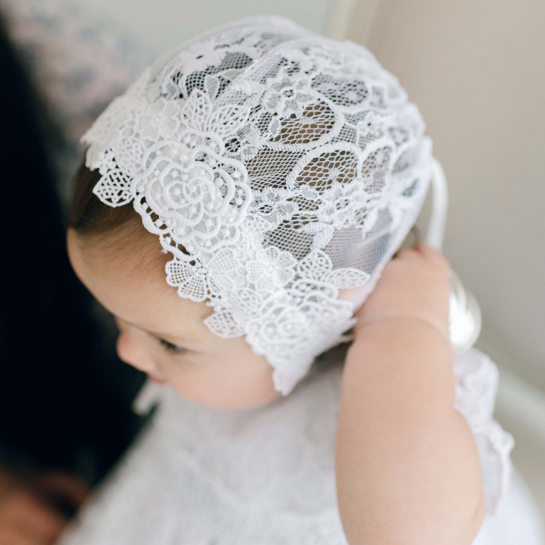 A baby, wearing an exquisite Olivia Lace Christening Bonnet, adjusts it with one tiny hand. Dressed in a white outfit that perfectly complements the detailed lace pattern of the bonnet, the scene is charmingly set against a softly blurred background.