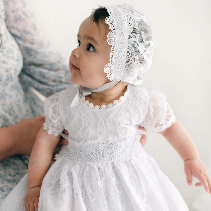 A baby wearing a white Christening gown paired with the Olivia Lace Christening Bonnet looks to the side, while an adult's hand gently supports them. The softly blurred background enhances the intricate lace details of the outfit.