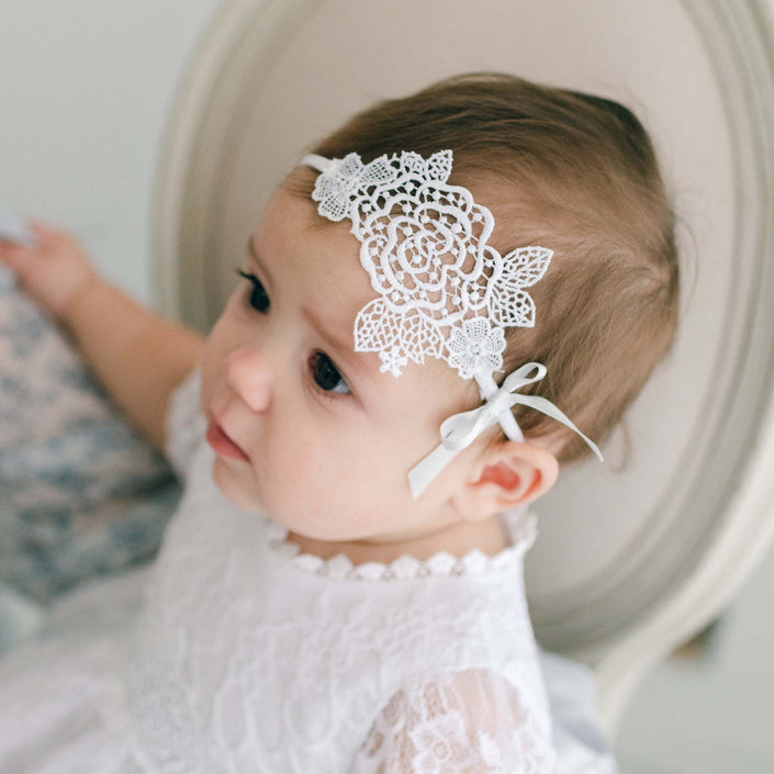 A baby wearing a white rose lace dress sits on a chair, sporting an Olivia Lace Headband handmade with a floral pattern and ribbon tied to the side. With a neutral expression, the baby glances off to the side.