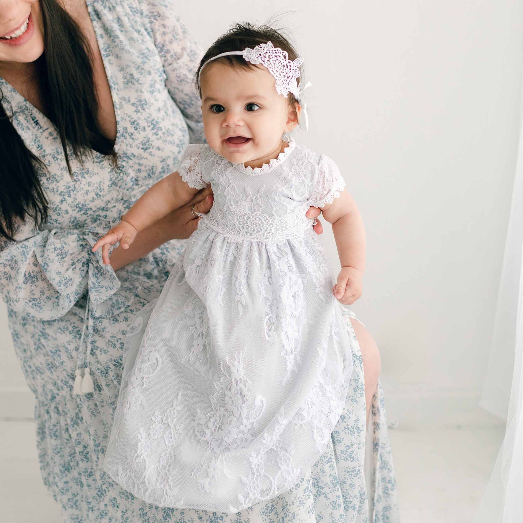A woman cradles a smiling baby dressed in a delicate Olivia Layette gown, complete with a matching headband. The woman wears a floral-patterned blouse, and the softly lit background is bathed in gentle white tones.