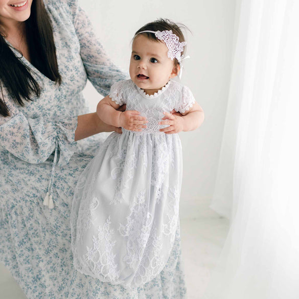 In a softly lit room with sheer white curtains in the background, a baby dressed in an Olivia Layette, paired with a matching headband, is held by a woman wearing a coordinating floral-patterned gown.