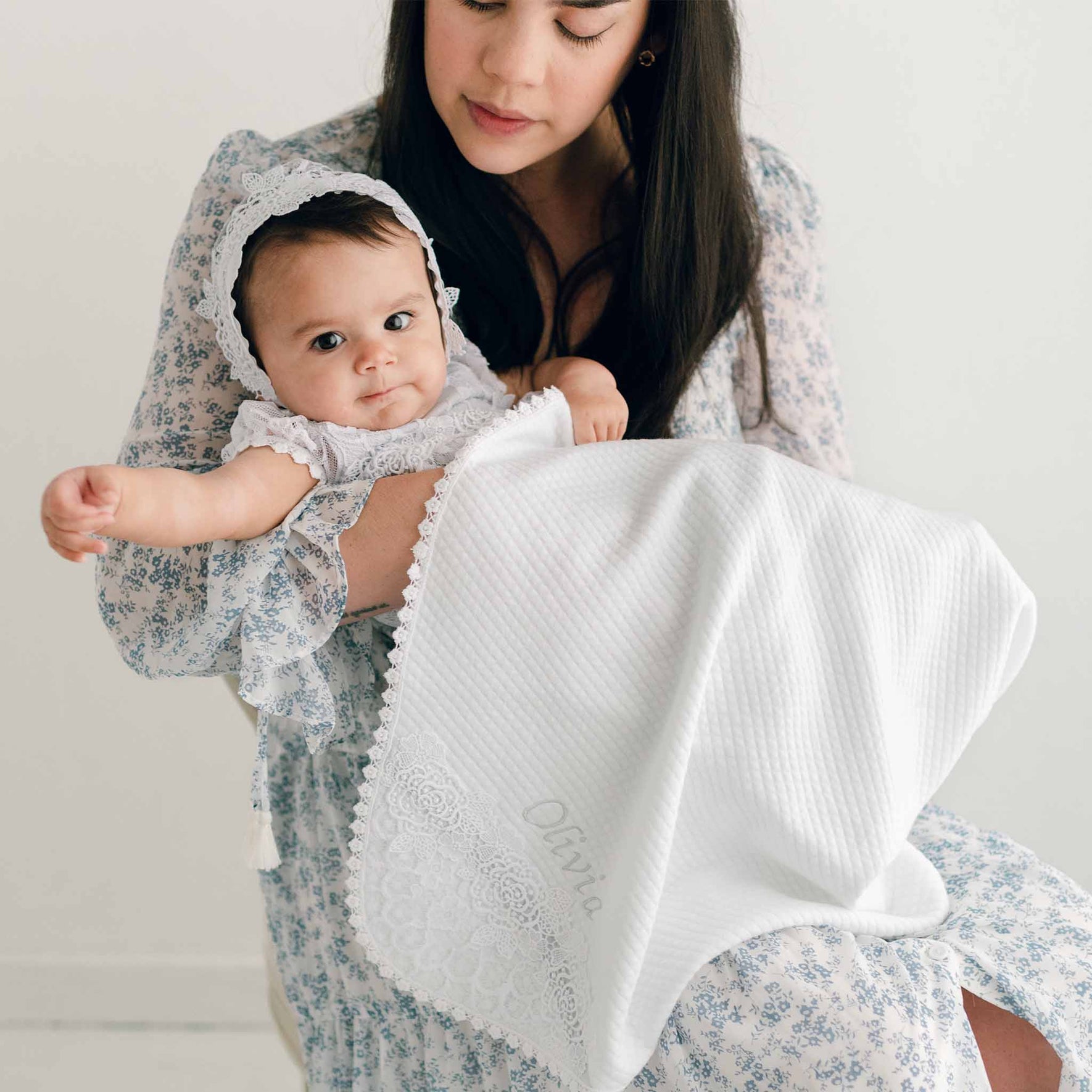 A woman in a floral dress holds a baby wearing a bonnet and patterned dress, wrapped in the Olivia Personalized Blanket, which features embroidered details for an added personalized touch. The background is plain and softly lit.
