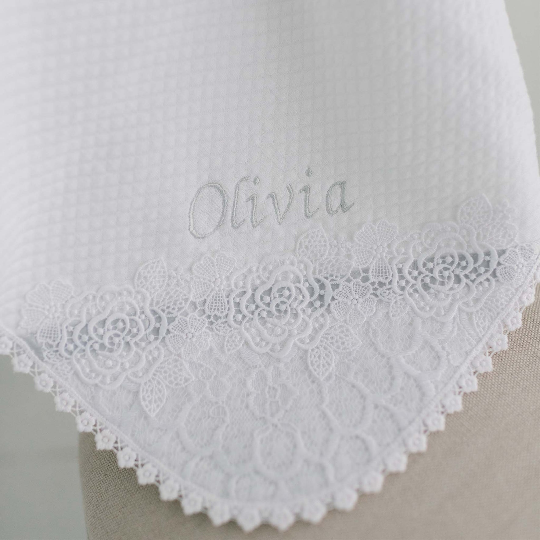 A close-up of the Olivia Personalized Blanket highlights the name "Olivia" elegantly embroidered on white fabric. The waffle texture is beautifully complemented by a floral lace trim, adding a personalized touch with its intricate patterns along the edge.