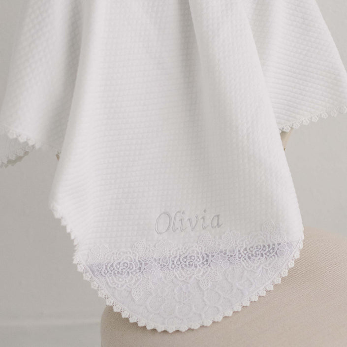 A white fabric from the Olivia Personalized Blanket drapes over the edge of a chair, featuring intricate floral lace detailing along the border. The name "Olivia" is embroidered in light thread near the center, offering a personalized touch reminiscent of a cherished Christening blanket.