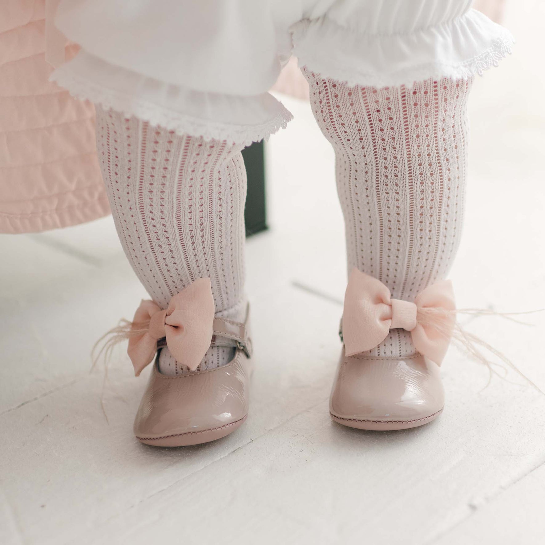 A close-up photo captures a child's legs adorned with Openwork Fancy Tights featuring an intricate white lace pattern, light pink shoes decorated with bows, and white ruffled pants. The child is standing on a light-colored floor.
