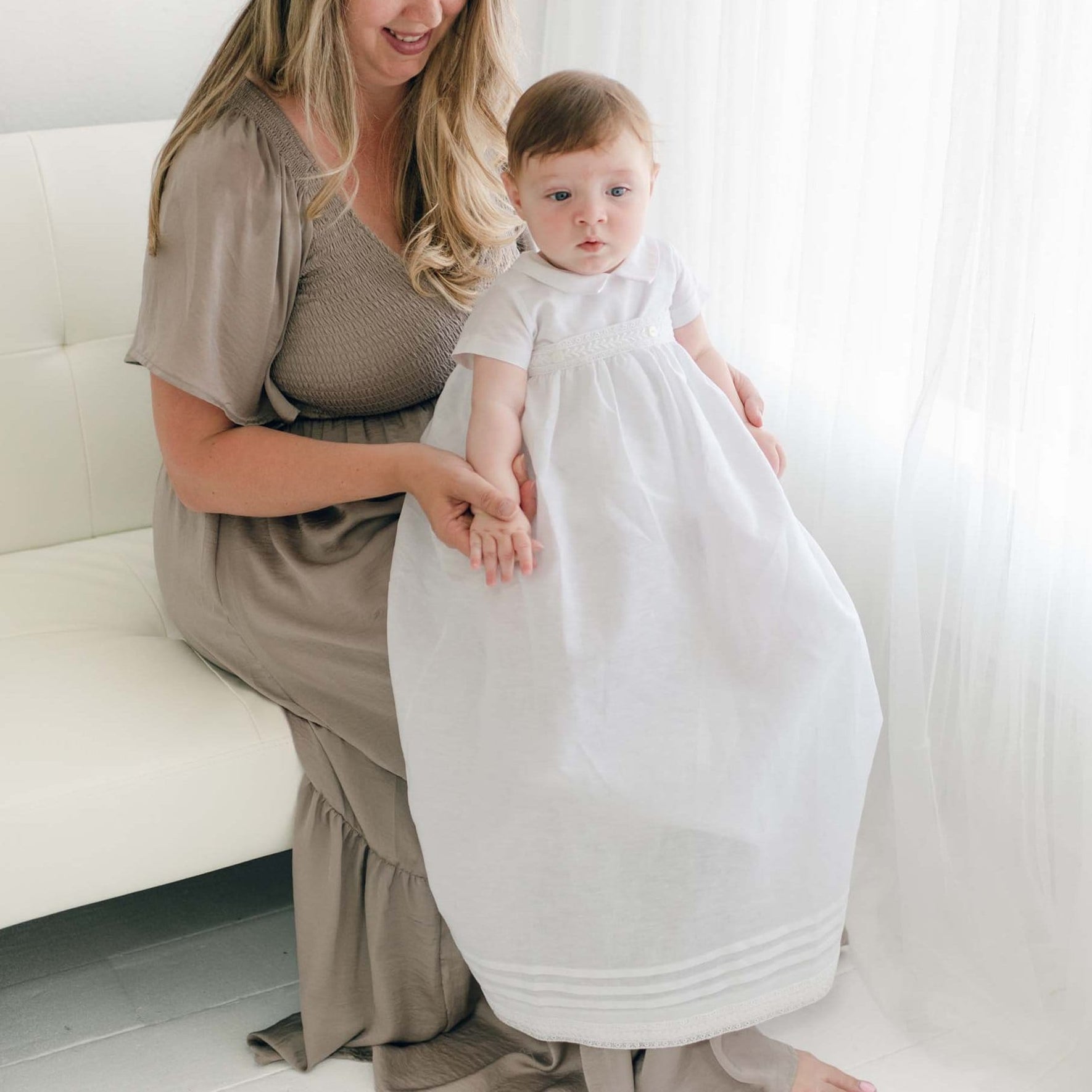 A woman with long hair in a brown dress sits on a white couch, holding a baby in an Oliver Convertible Gown | Romper & Skirt Set. They are both facing the camera near a bright window with white curtains.