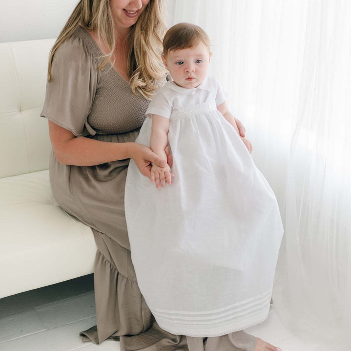 A woman with long hair in a brown dress sits on a white couch, holding a baby in an Oliver Convertible Gown | Romper & Skirt Set. They are both facing the camera near a bright window with white curtains.