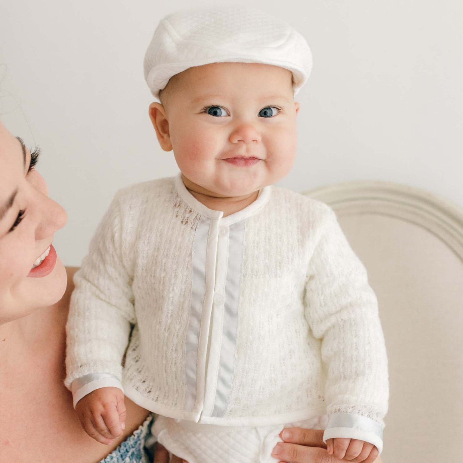 Baby boy smiling with his mother and wearing the Owen Knit Sweater made from a soft knit in ivory and trimmed with ivory linen and blue silk ribbon