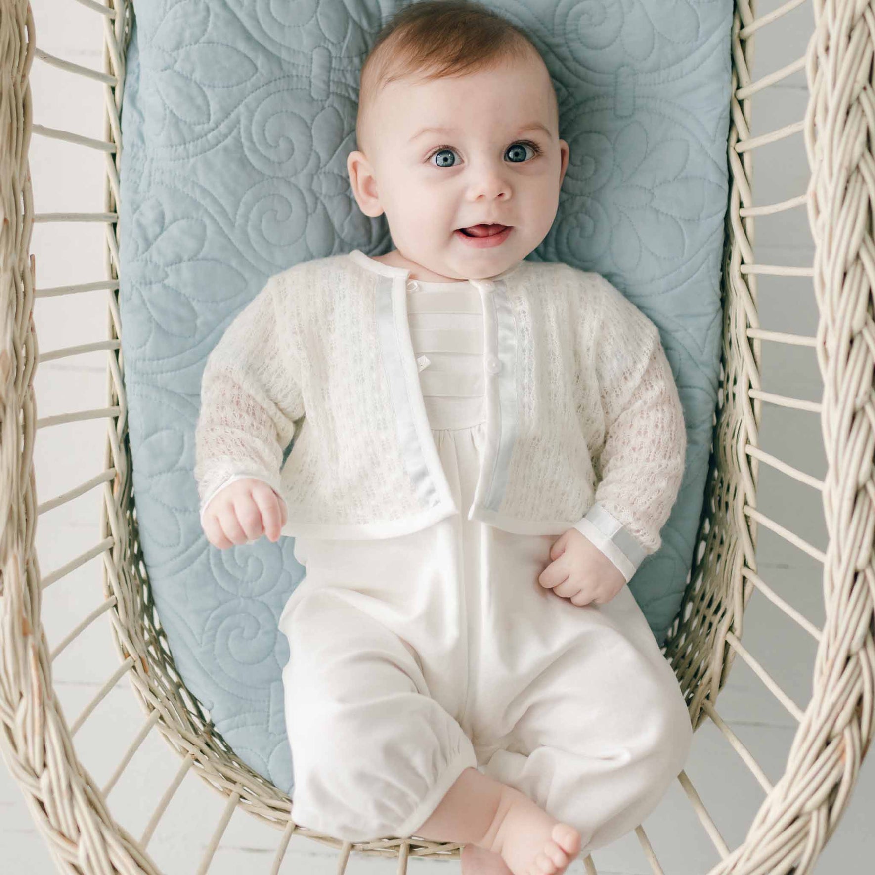 A baby with light hair and blue eyes is sitting in a wicker chair lined with a quilted cushion. The baby, dressed in a white Christening outfit and an Owen Knit Christening Sweater handmade in the USA, is smiling while looking slightly to the side.