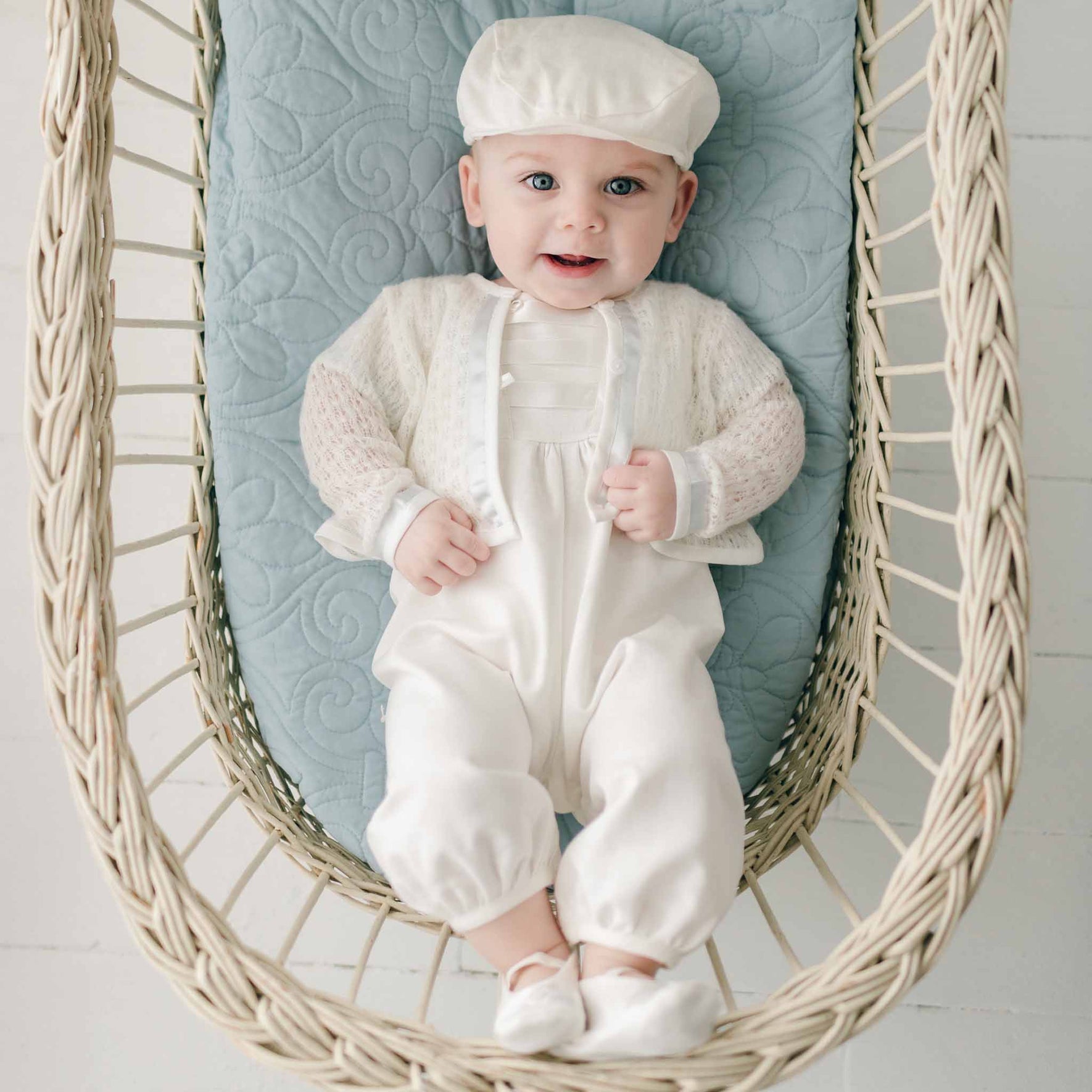A baby wearing an Owen Knit Christening Sweater and hat is lying on a light blue quilt inside a woven basket. The baby is looking up with a smile, hands resting on the chest and feet comfortably laid out in the basket. The charming scene, handmade in the USA, features a basket on a white wooden floor.