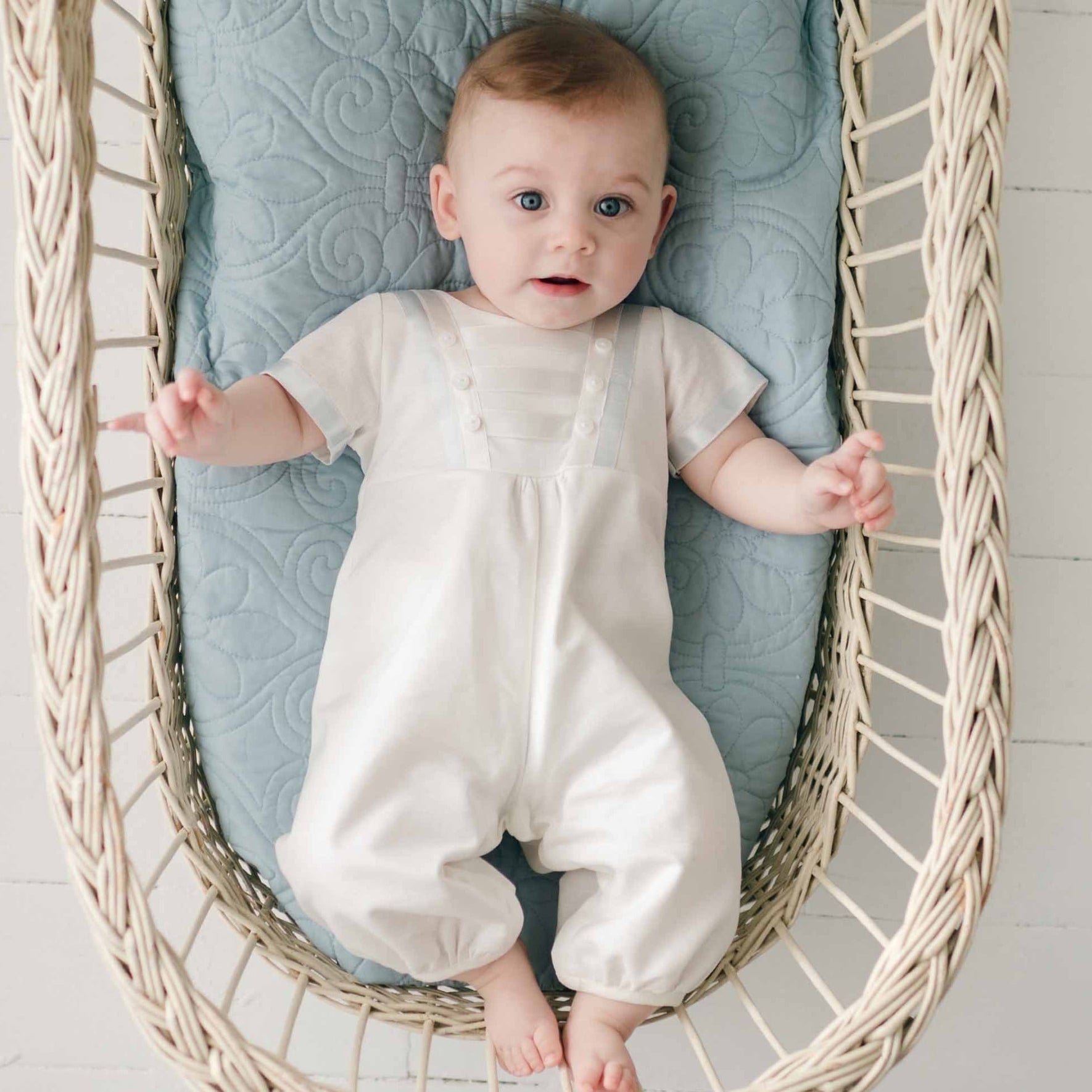A baby with light hair, clad in an Owen Linen Christening Romper, is lying on a blue quilted cushion in a woven bassinet. The baby is looking up with arms stretched out to the sides. The background is a white surface.