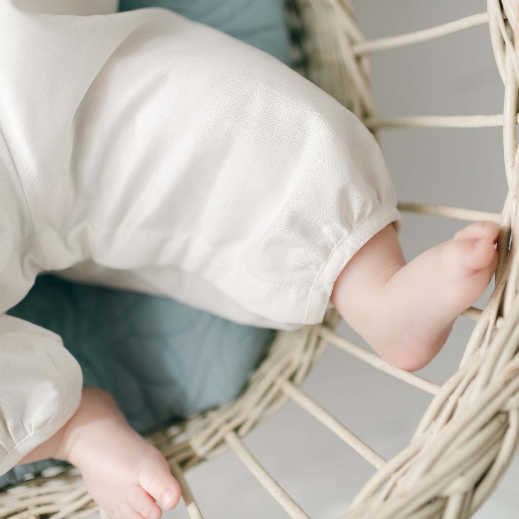 Close-up image of a baby’s legs and feet wearing an Owen Linen Christening Romper, sitting in a white wicker chair. The baby is resting on a blue cushion. Only the lower part of the baby's body is visible.