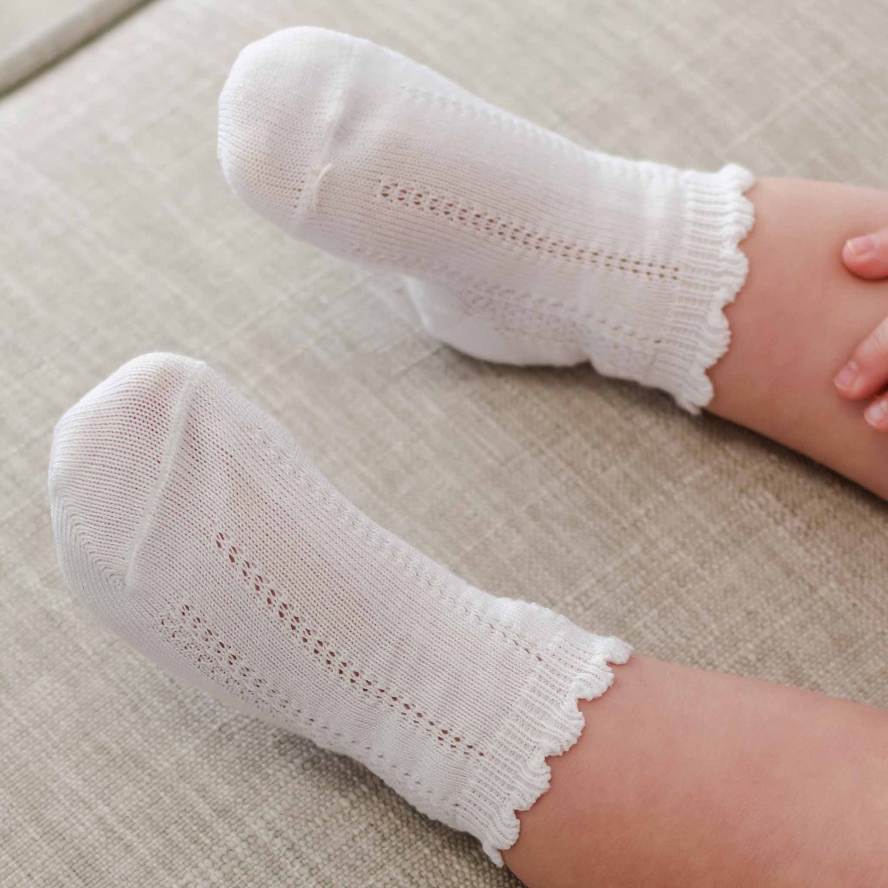 Close-up of a child's feet in Pattern Socks, crafted from white cotton with a delicate knit design and scalloped cuffs. The child is seated on a light-colored fabric surface.