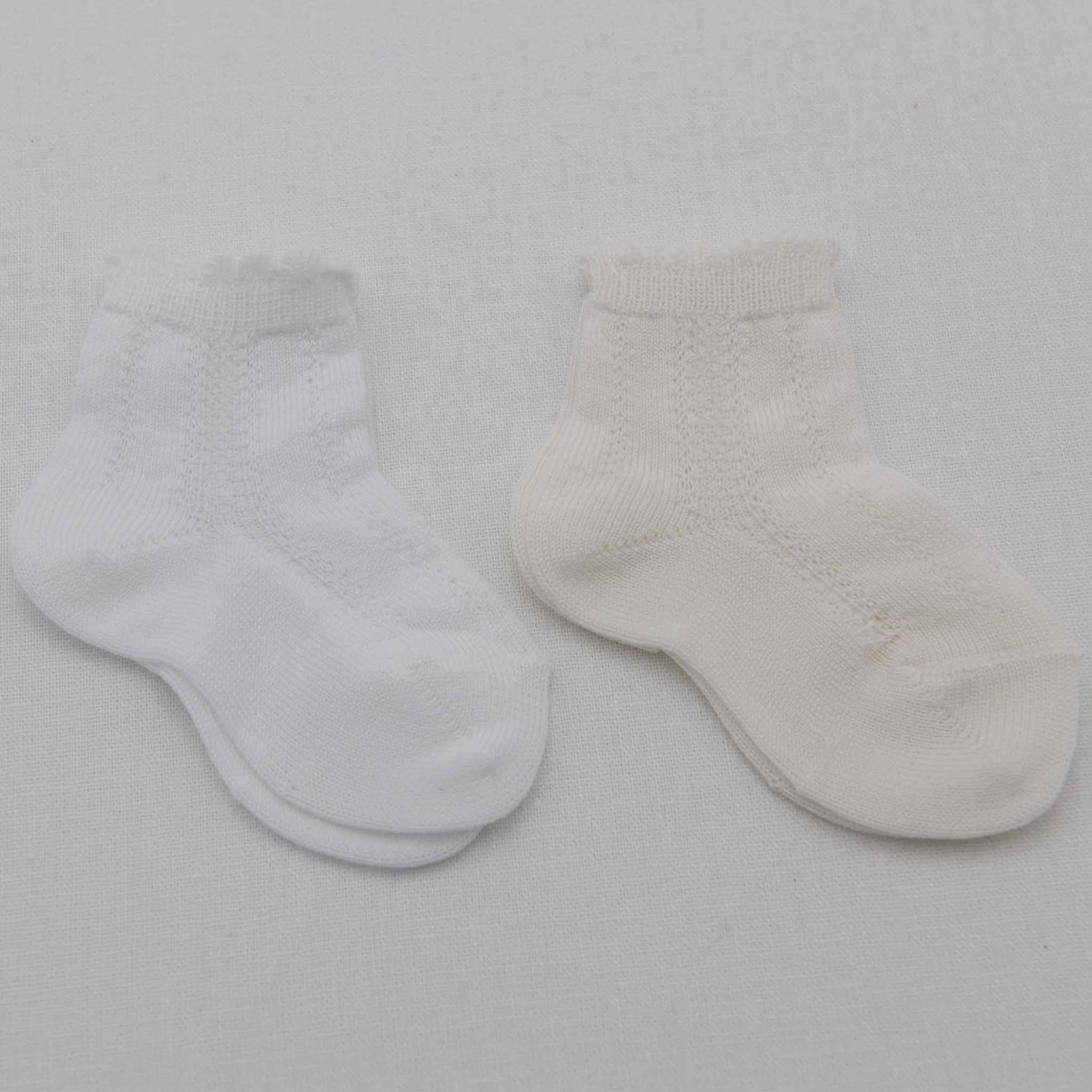 A pair of Pattern Socks, designed for babies in a small size, are placed next to each other on a plain white surface. The sock on the left is white, while the one on the right has a light cream shade, each featuring a subtle knit pattern. They are crafted from soft cotton and adorned with delicate scalloped cuffs that add a charming touch.