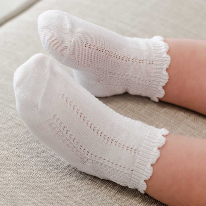 Tiny feet adorned in white "Pattern Socks" rest on a light-colored surface. These socks showcase delicate knit designs and a charming scalloped cuff along the top.