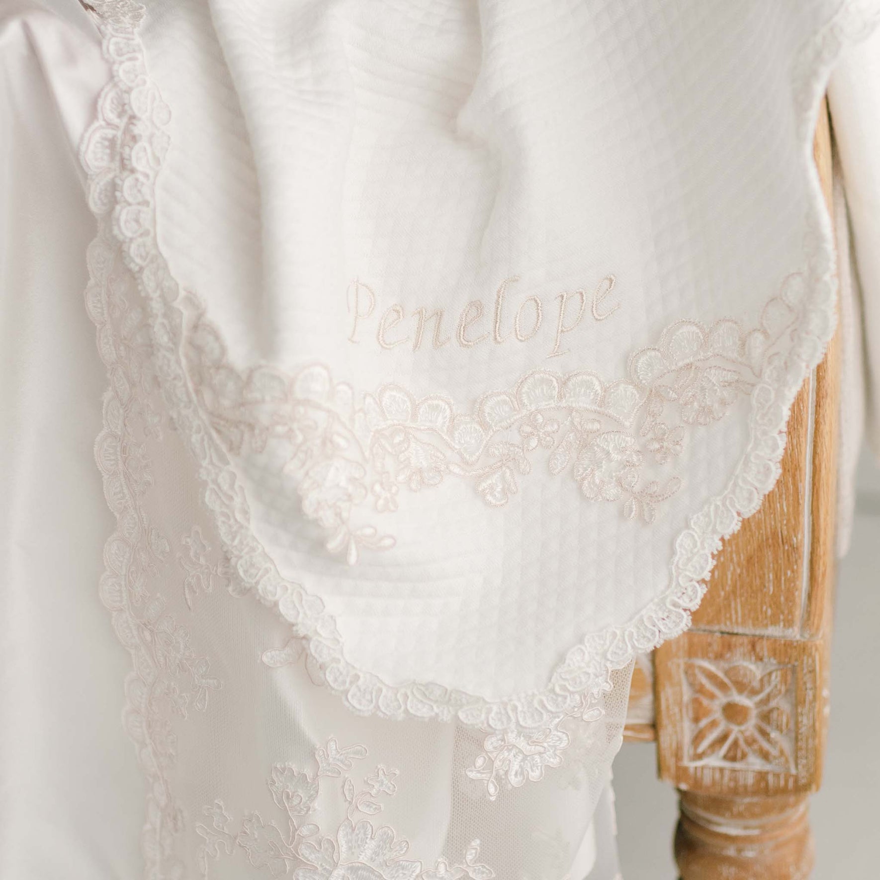 Personalized baptism blanket draped over chair, baby girl name Penelope embroidered in silk on corner.