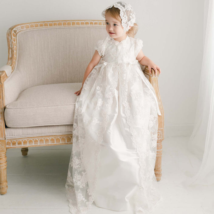 Baby girl sitting in chair wearing the Penelope heirloom Christening Gown made of luxurious silk and lace netting. 