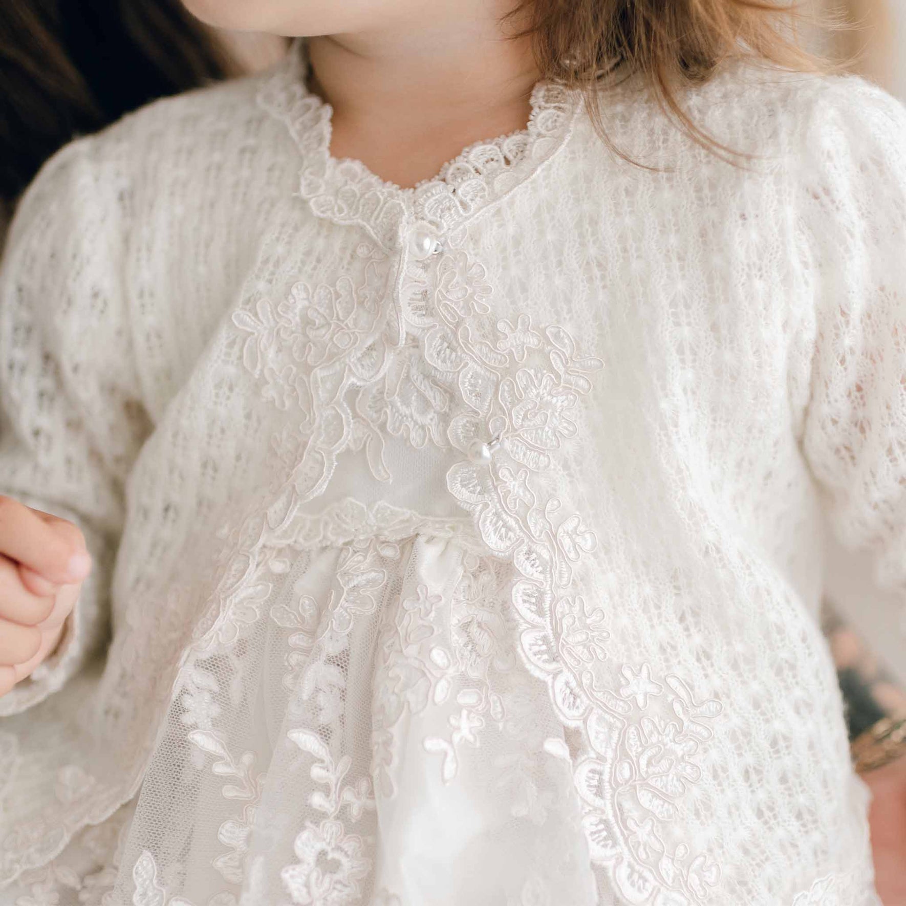 A child is dressed in a white, meticulously crafted article from the Penelope Knit Bonnet & Sweater collection, embellished with pearl-style buttons. This ensemble showcases floral embroidery and lace designs, featuring a scalloped neckline and long sleeves, emphasizing the texture and intricate design.