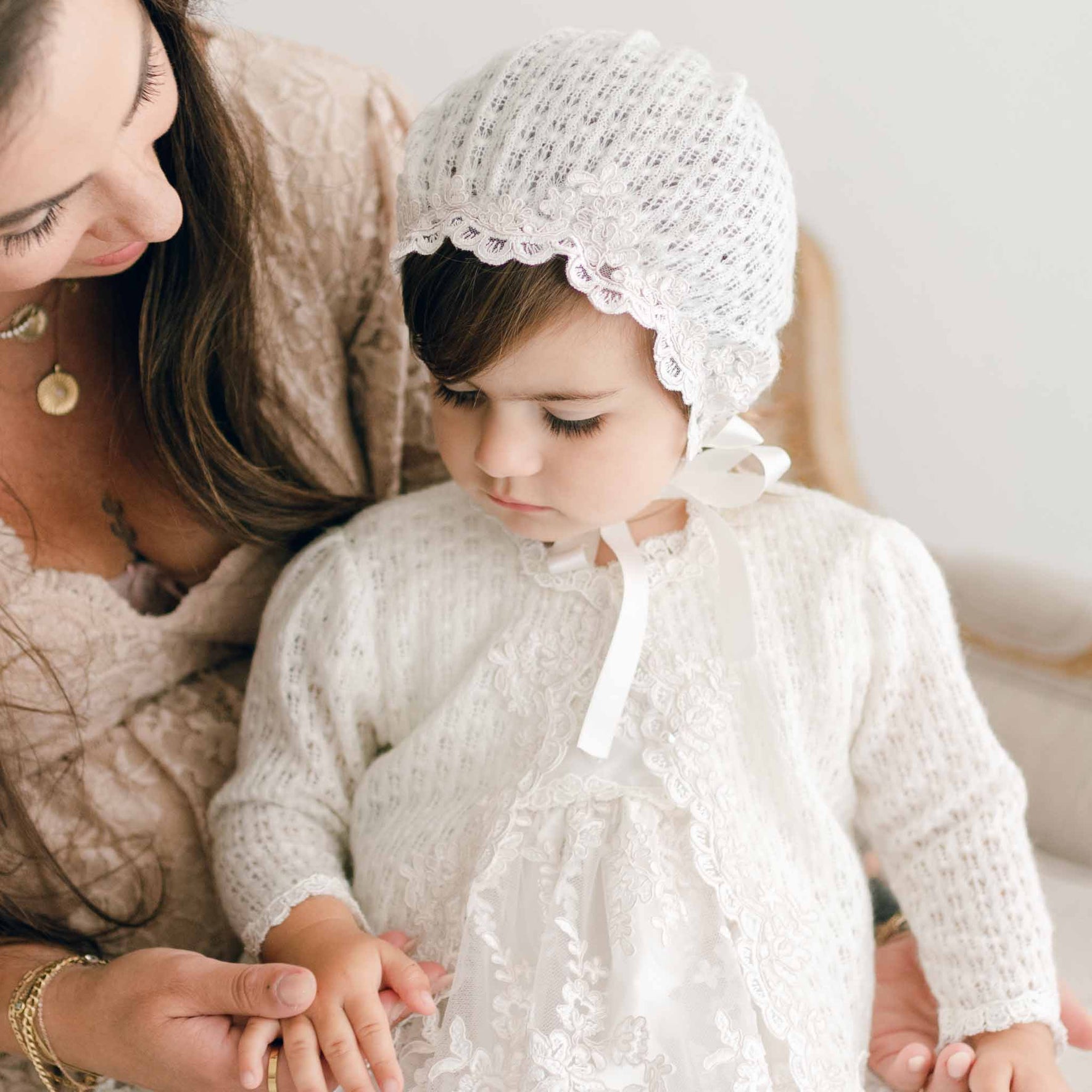 Baby girl looking at her hand with mom wearing her knit bonnet and sweater part of the Penelope Christening Collection.