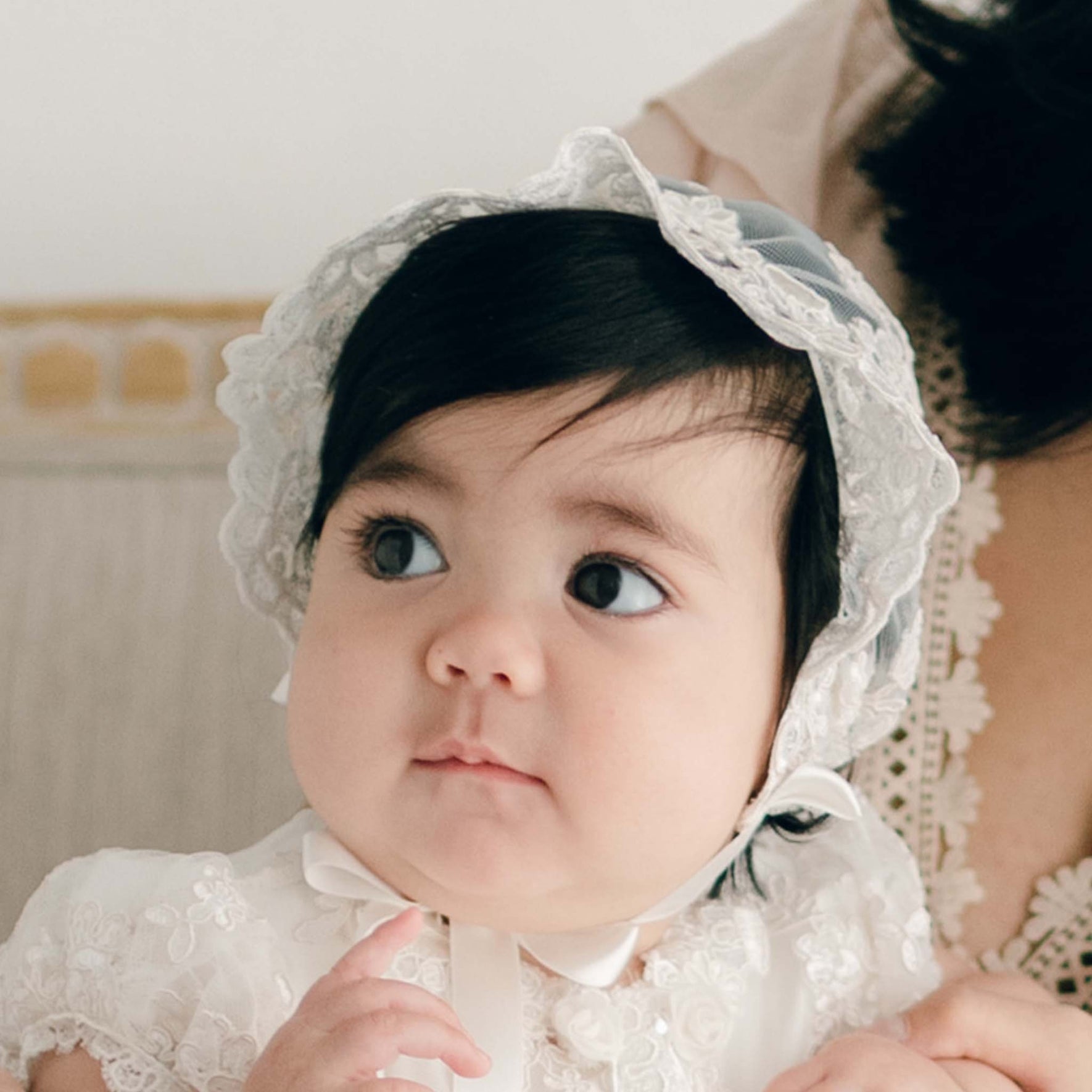 Baby girl wearing the Penelope Lace Christening Bonnet and matching gown sits on the lap of her mother.