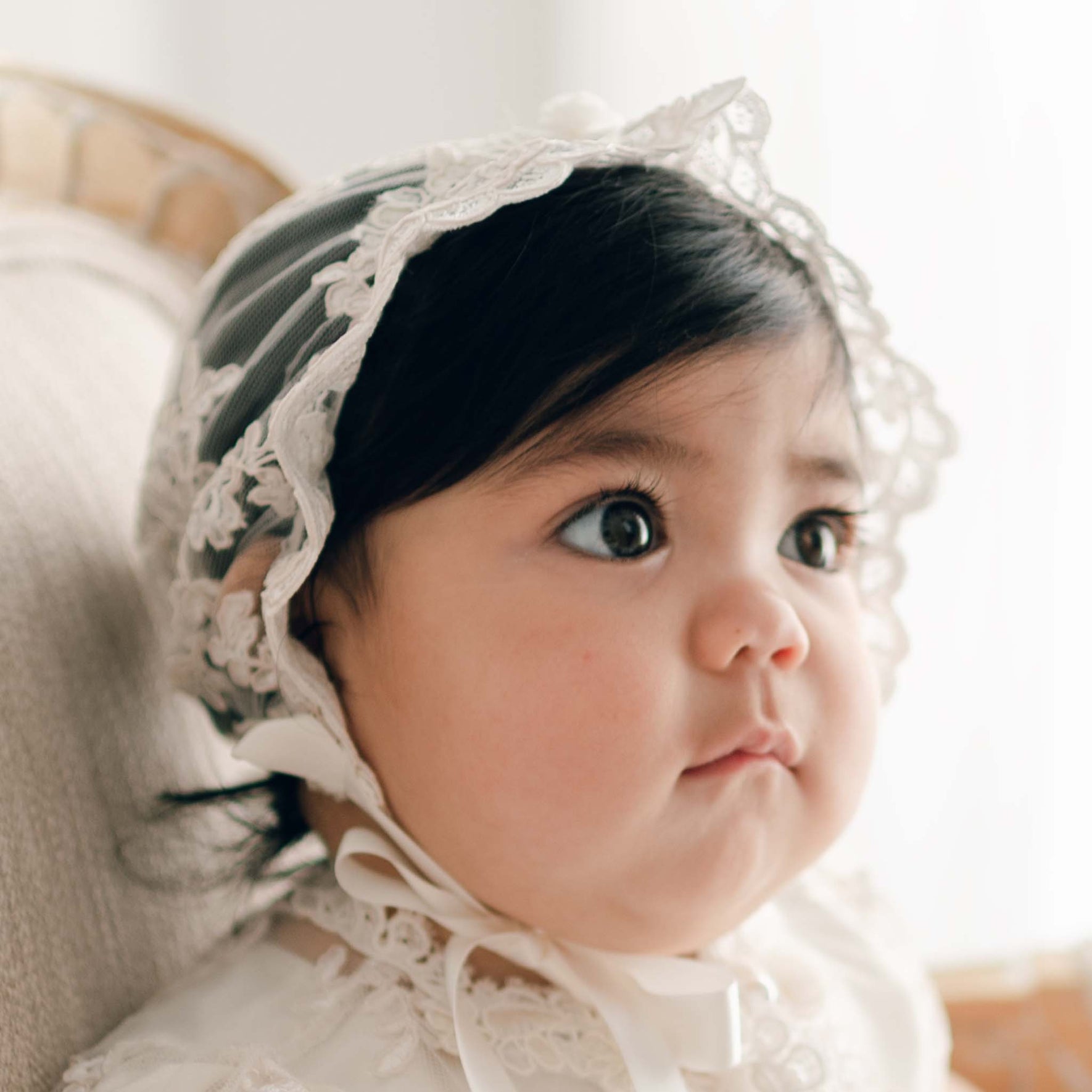 Baby girl wearing the Penelope Lace Christening Bonnet and matching baptism dress gazes away from the camera. 
