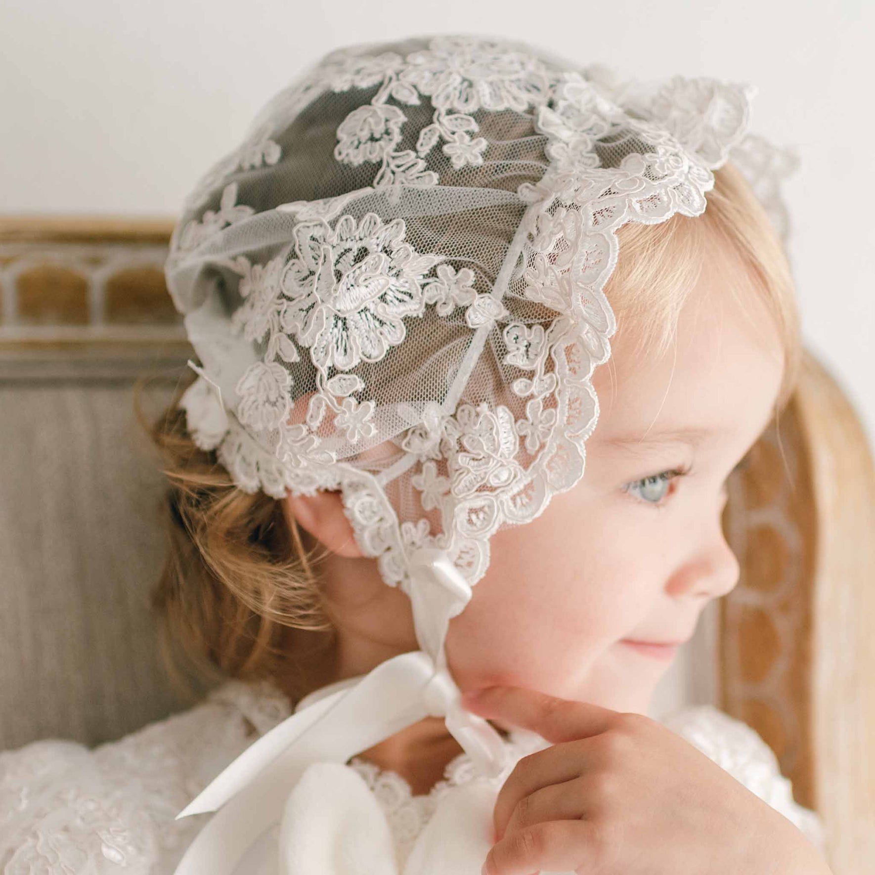 Close up detail of a baby girl smiling in profile sitting in a beige upholstered chair and wearing the Penelope Lace Christening Bonnet and matching gown.