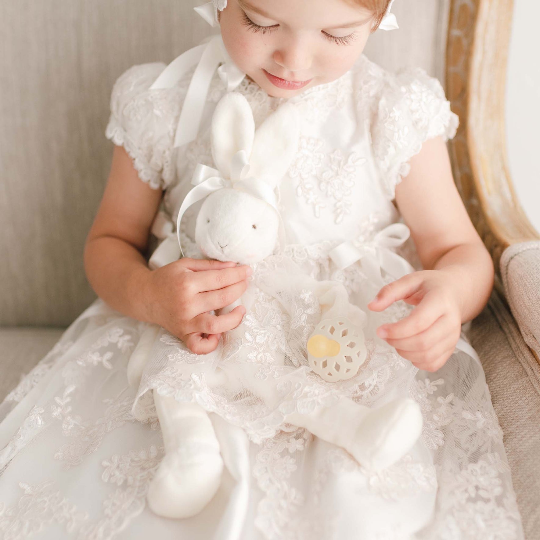 Close up photo of the Penelope silly bunny buddy doll. Doll is holding a rubber pacifier while wearing a christening dress.