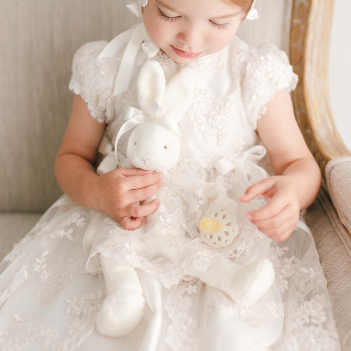 Close up photo of the Penelope silly bunny buddy doll. Doll is holding a rubber pacifier while wearing a christening dress.