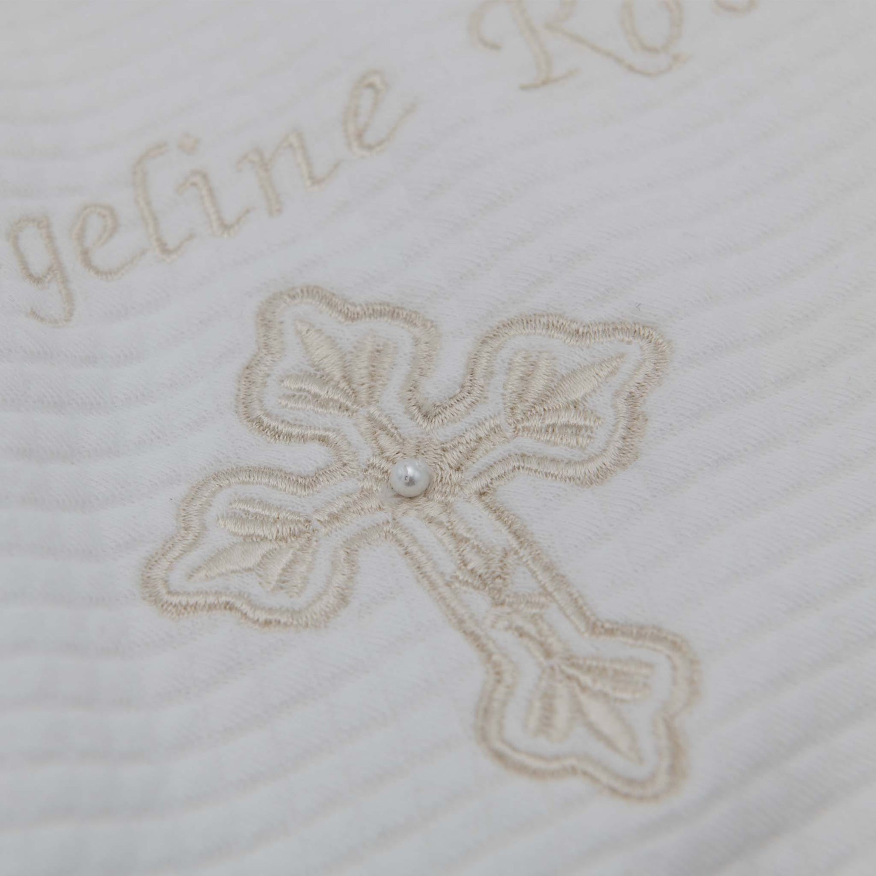 A detailed view of the Personalized Cross Blanket reveals its handmade design in textured cotton, featuring a white quilted fabric with an embroidered beige cross adorned by a small pearl at the center. Above the cross, an elegantly embroidered name in beige script adds a personal touch.