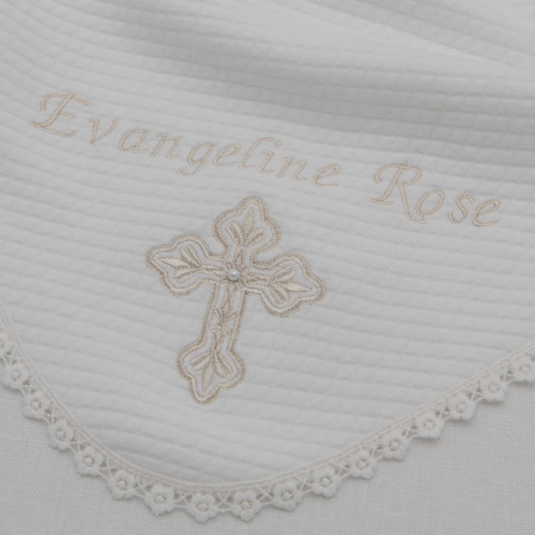 The Personalized Cross Blanket showcases white quilted fabric with the name "Evangeline Rose" elegantly embroidered in cursive. Below the name, a floral cross design is artfully stitched, and the blanket's textured cotton edge is embellished with delicate lace trim, highlighting fine artisanal craftsmanship.