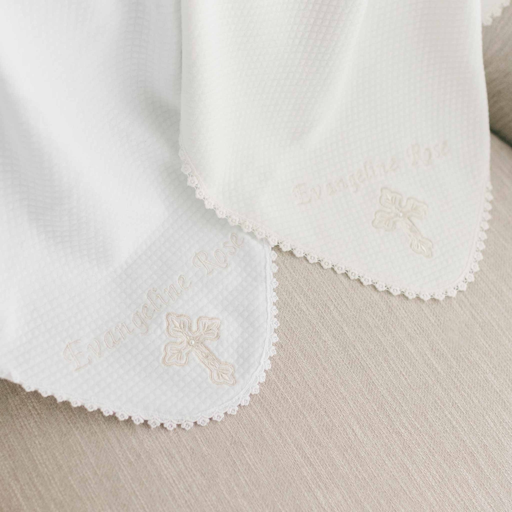 A close-up image reveals two white Personalized Cross Blankets with scalloped edges, each delicately embroidered with a cross and the name "Evangeline Rose," resting on a light-colored couch. The blankets display a subtle waffle pattern, enhancing their textured cotton appearance.