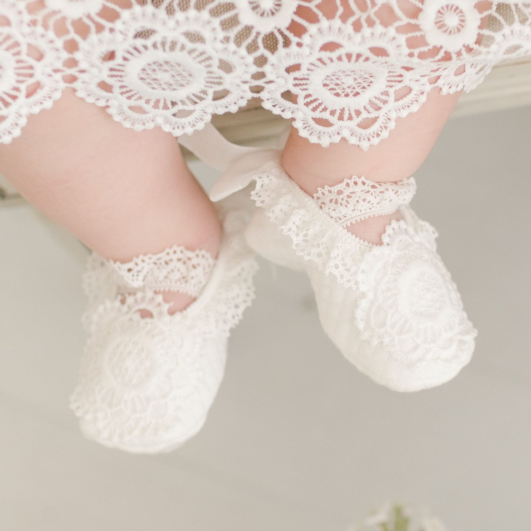 Close-up of the Poppy Christening Booties adorned with delicate floral lace and soft white fabric, peeking from under the detailed lace hem of the Poppy Blessing Dress.