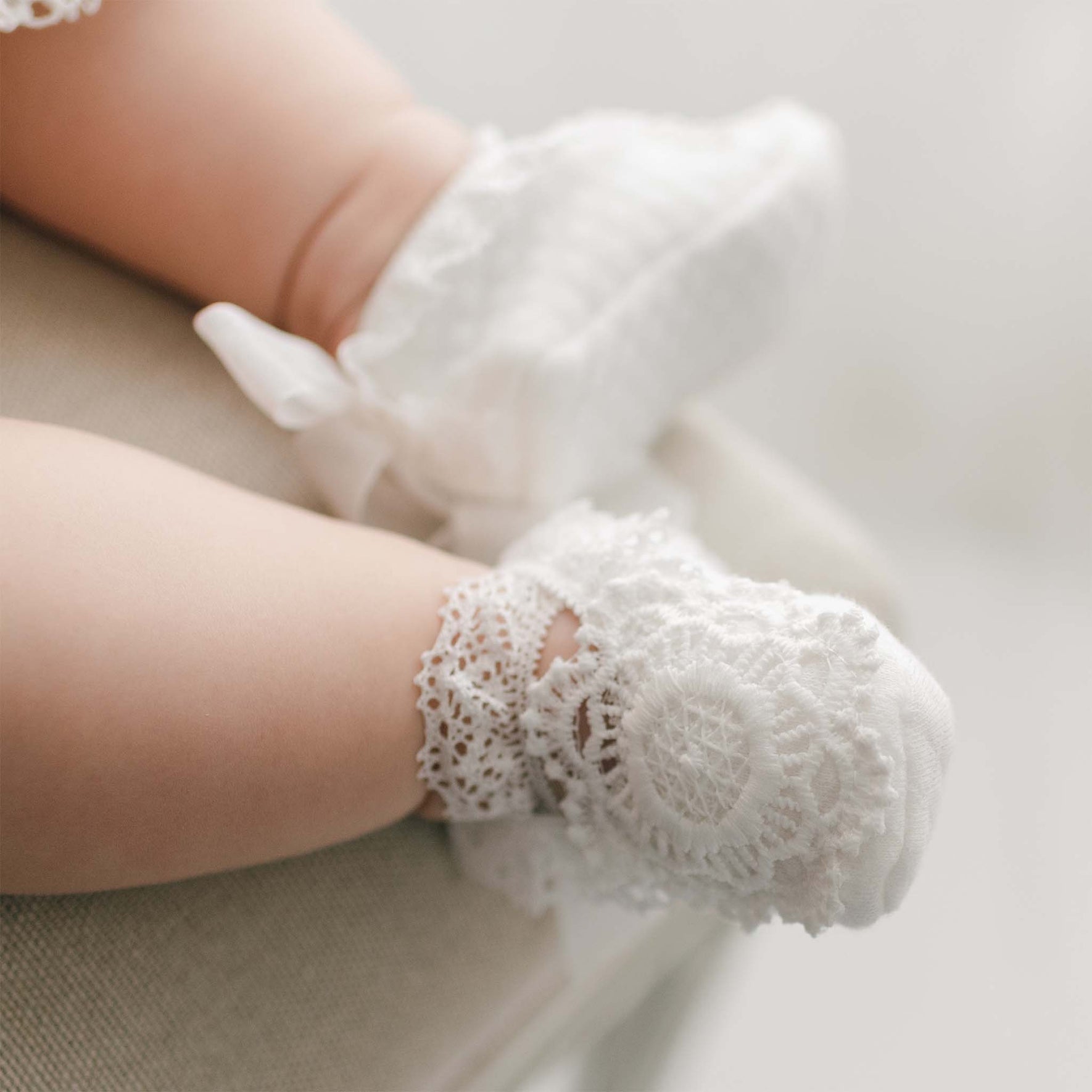 Detailed view of the Poppy Christening Booties on baby feet, showcasing intricate lace details and white ribbon accents, placed softly on a neutral fabric background.